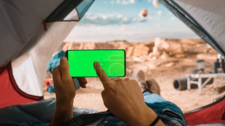 Photo for POV View of a Tourist Horizontally Holding a Smartphone with Green Screen Placeholder, Tapping a Finger on Display. Traveller Resting in a Tent on Top of a Rocky Mountain and Flying Hot Air Balloons. - Royalty Free Image