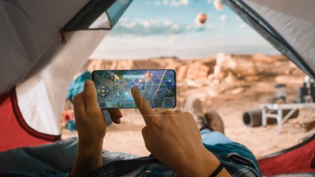 POV eines Touristen, der ein Smartphone benutzt, um ein Arcade-Handyspiel zu spielen, wischt mit dem Finger über das Display. Reisende ruhen sich in einem Zelt auf einem felsigen Berg aus und fliegen Heißluftballons.