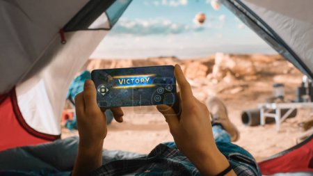 Photo for POV of a Tourist Using Smartphone For Playing Arcade Mobile Game, Celebrating Victory after successful Round. Traveller Resting in a Tent on Top of a Rocky Mountain and Flying Hot Air Balloons. - Royalty Free Image