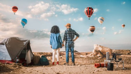 Photo for Young Diverse Tourist Couple Hiking and Living in a Tent in Great Outdoors in Rocky Canyon Valley. Male and Female Backpacker Hold Hands and Look at Hot Air Balloon Festival in Mountain National Park. - Royalty Free Image