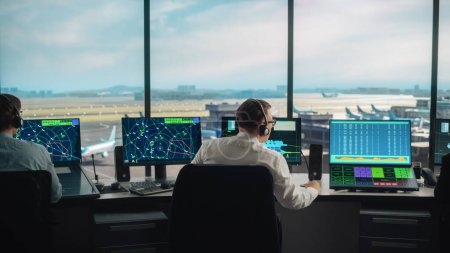 Diverse Fluglotsen arbeiten in einem modernen Flughafen-Tower. Der Büroraum ist voll mit Desktop-Computer-Displays mit Navigationsbildschirmen, Abflug- und Ankunftsdaten für Fluglotsen.