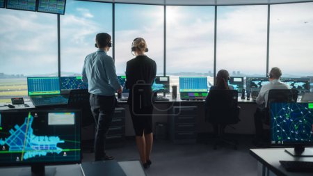 Weibliche und männliche Fluglotsen mit Headsets unterhalten sich im Flughafen-Tower. Der Büroraum ist voll mit Desktop-Computer-Displays mit Navigationsbildschirmen, Abflug- und Ankunftsdaten des Flugzeugs für das Team.