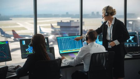 Controladores de Tráfico Aéreo Femenino y Masculino con Auriculares Talk in Airport Tower. La sala de oficina está llena de pantallas de computadora de escritorio con pantallas de navegación, salidas de aviones y datos de llegada para el equipo.