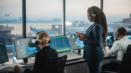 Schwarze Fluglotsin hält Tablet im Flughafen-Tower Der Büroraum ist voll mit Desktop-Computer-Displays mit Navigationsbildschirmen, Abflug- und Ankunftsdaten des Flugzeugs für das Team.