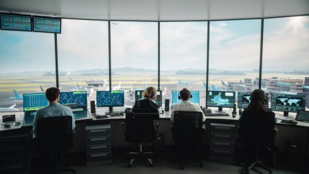 Diverse Fluglotsen arbeiten in einem modernen Flughafen-Tower. Der Büroraum ist voll mit Desktop-Computer-Displays mit Navigationsbildschirmen, Abflug- und Ankunftsdaten für Fluglotsen.