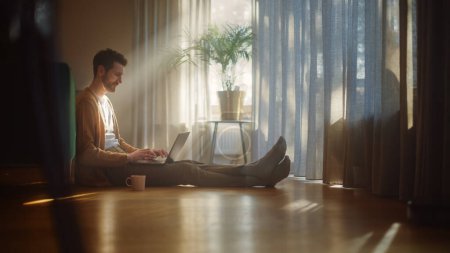 Photo for Handsome Happy Man Using Laptop at Home for Remote Work. Early Middle Aged Smiling Guy Sitting on the Floor Uses Internet, Writes Report, Finishes Project. Good Sunny Day for Productive Distant Work - Royalty Free Image