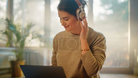 Téléchargez les photos : Young Woman Using Laptop at Home, Does Remote Work, Écoute de la musique à travers des écouteurs. Belle fille souriante assise sur le sol aime la musique, danse un peu, remue-méninges projet créatif - en image libre de droit