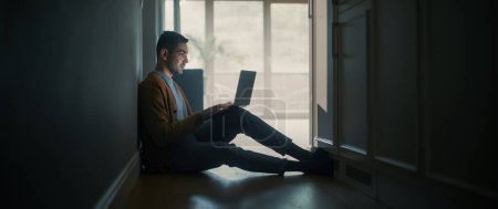 Téléchargez les photos : Portrait d'un jeune homme adulte utilisant un ordinateur portable à la maison. Il est assis sur le sol du couloir, dos au mur. Utilise Internet pour le travail à distance productif, Divertissement sur les médias sociaux. - en image libre de droit