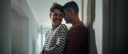 Foto de Escenas suaves de una pareja gay de adultos jóvenes con estilo pasan tiempo en casa. Dos hombres felices enamorados se paran en el pasillo con ropa casual y se abrazan. Lindo contenido de relación LGBT. - Imagen libre de derechos