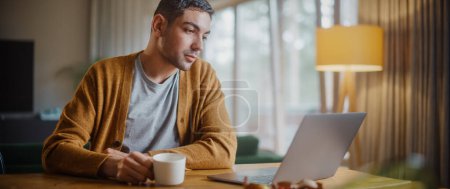 Photo for Handsome Adult Man Using Laptop Computer, Sitting in Living Room and Drinking Tea or Coffee in Apartment. Attractive Man is Online Shopping on Internet, Watching Funny Videos on Streaming Service. - Royalty Free Image