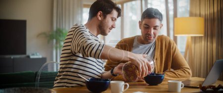 Photo for Two Handsome Friends are Eating Colorful Breakfast Cereal in Cozy Kitchen in Stylish Apartment. Young Adult Gay Couple Have a Conversation While Eating Healthy Nutricious Food in the Morning. - Royalty Free Image