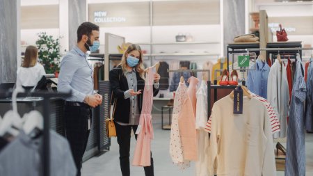 Photo for Clothing Store: Female Customer Shopping, Retail Sales Assistant Helps. Wearing Protective Face Masks. People in Fashionable Shop, Choosing Stylish Clothes, Colorful Brand with Sustainable Designs - Royalty Free Image