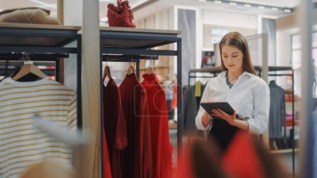 Photo for Clothing Store: Female Visual Merchandising Professional Uses Tablet Computer To Create Stylish Collection. Fashionable Shop Sales Retail Assistant Checks Stock. Small Business Owner Orders Items - Royalty Free Image