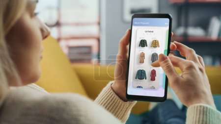 Beautiful Caucasian Female is Using Smartphone with Clothing Online Web Store to Choose and Buy Clothes from New Collection. Female Surfing the Net and Lying on Couch Sofa at Home Living Room.