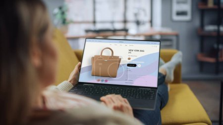 Beautiful Caucasian Female is Using Laptop Computer with Clothing Online Web Store to Choose and Buy Handbag from New Collection. Female Surfing the Net and Lying on Couch Sofa at Home Living Room.