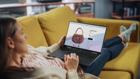 Hermosa hembra caucásica está utilizando el ordenador portátil con ropa en línea Web Store para elegir y comprar bolso de la nueva colección. Mujer navegando por la red y acostada en el sofá sofá en la sala de estar en casa.