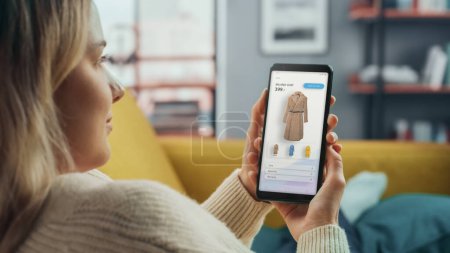 Beautiful Caucasian Female is Using Smartphone with Clothing Online Web Store to Choose and Buy Coat from New Collection. Female Surfing the Net and Lying on Couch Sofa at Home Living Room.