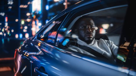 Téléchargez les photos : Élégant homme noir à lunettes est de retour à la maison dans un siège arrière d'un taxi la nuit. Beau passager masculin regardant par la fenêtre tandis que dans une voiture dans la rue de la ville urbaine avec des panneaux au néon de travail. - en image libre de droit