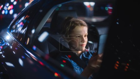 Schöner kleiner Junge sitzt auf dem Rücksitz eines Autos und pendelt nachts nach Hause. Fahrgast spielt Videospiel auf Tablet-Computer im Taxi in der City Street mit funktionierenden Leuchtreklamen.