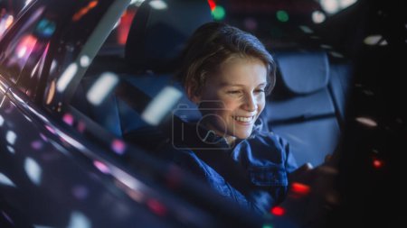 Téléchargez les photos : Young Boy est assis sur le siège arrière d'une voiture, se rendant à la maison la nuit. Regarder les passagers Divertissement drôle sur Smartphone tout en taxi dans la rue de la ville avec des panneaux au néon de travail. - en image libre de droit