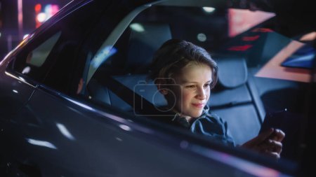 Photo for Young Boy is Sitting on Backseat of a Car, Commuting Home at Night. Passenger Watching Funny Entertainment on Smartphone while in Taxi in City Street with Working Neon Signs. - Royalty Free Image