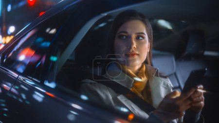 Photo for Stylish Female is Commuting Home in a Backseat of a Taxi at Night. Beautiful Woman Passenger Using Smartphone and Looking Out of Window while in a Car in Urban City Street with Working Neon Signs. - Royalty Free Image