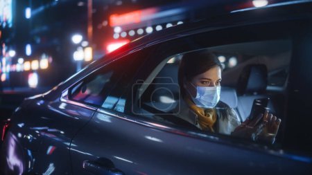 Mujer usando máscara facial es conmutar a casa en el asiento trasero de un taxi por la noche. Hermoso pasajero usando Smartphone mientras está en un coche en Urban City Street con letreros de neón de trabajo.