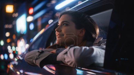 Téléchargez les photos : La jeune femme excitée est assise sur le siège arrière d'une voiture, faisant la navette vers la maison la nuit. Regardant par la fenêtre avec étonnement de la beauté de la rue de la ville avec des panneaux au néon qui fonctionnent. - en image libre de droit