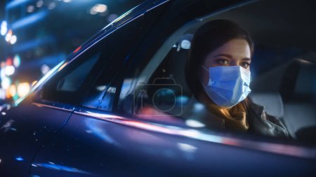 Mujer usando máscara facial es conmutar a casa en el asiento trasero de un taxi por la noche. Hermoso pasajero mirando por la ventana mientras está en un coche en Urban City Street con letreros de neón de trabajo.