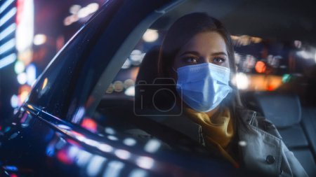 Mujer usando máscara facial es conmutar a casa en el asiento trasero de un taxi por la noche. Hermosa pasajera mirando por la ventana mientras está en un coche en Urban City Street con letreros de neón de trabajo.