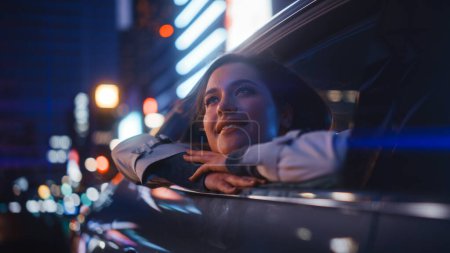 Téléchargez les photos : La jeune femme excitée est assise sur le siège arrière d'une voiture, faisant la navette vers la maison la nuit. Regardant par la fenêtre avec étonnement de la beauté de la rue de la ville avec des panneaux au néon qui fonctionnent. - en image libre de droit