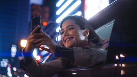 Photo for Excited Female is Looking Out of the Window from Backseat of a Car at Night. Woman Taking Photos and Videos on a Smartphone with Amazement of How Beautiful is the City Street with Working Neon Signs. - Royalty Free Image