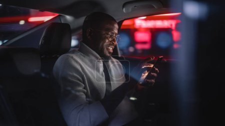 Foto de Happy Black Man in Glasses está viajando a casa en un asiento trasero de Taxi por la noche. Hombre guapo usando Smartphone y sonriendo mientras está en un coche en Urban City Street con letreros de neón de trabajo. - Imagen libre de derechos