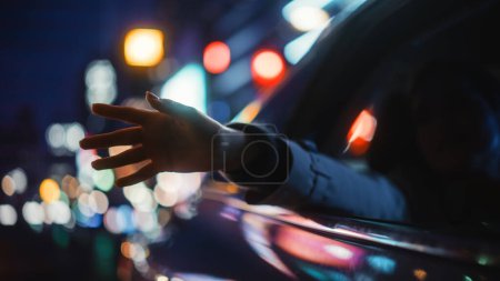Photo for Female is Commuting Home in a Backseat of a Taxi at Night. Passenger Chilling and Holding Her Hand Outside of Window while in a Car in Urban City Street with Working Neon Signs. - Royalty Free Image