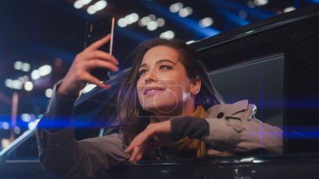 Photo for Excited Female is Looking Out of the Window from Backseat of a Car at Night. Woman Taking Photos and Videos on a Smartphone with Amazement of How Beautiful is the City Street with Working Neon Signs. - Royalty Free Image