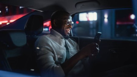 Foto de Elegante hombre negro en gafas está viajando a casa en un asiento trasero de un taxi por la noche. Hombre guapo haciendo una videollamada en un teléfono inteligente mientras está en el coche de transferencia en la calle de la ciudad con señales de neón de trabajo. - Imagen libre de derechos