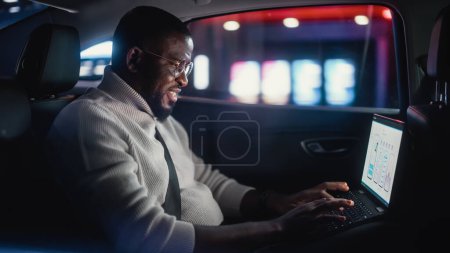 Stilvoller schwarzer Mann mit Brille pendelt nachts auf dem Rücksitz eines Taxis nach Hause. Mann benutzt Laptop und schaut aus dem Fenster, während er in einem Auto in der Stadtstraße mit Leuchtreklamen arbeitet.