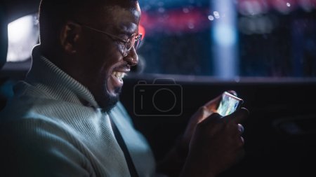 Photo for Stylish Black Man in Glasses is Commuting Home in a Backseat of a Taxi at Night. Handsome Male Playing Strategy Video Game on Smartphone while in a Car in Urban City Street with Working Neon Signs. - Royalty Free Image
