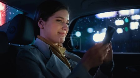 Stylish Female pendelt nachts auf dem Rücksitz eines Taxis nach Hause. Schöne Beifahrerin mit Smartphone und Blick aus dem Fenster im Auto in der Stadtstraße mit Leuchtreklamen.