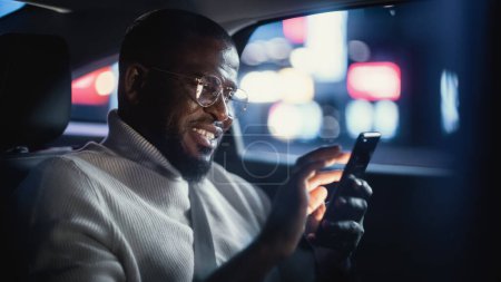 Photo for Happy Black Man in Glasses is Commuting Home in a Backseat of Taxi at Night. Handsome Male Using Smartphone and Smiling while in a Car in Urban City Street with Working Neon Signs. - Royalty Free Image