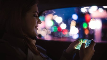 Photo for Happy Female is Commuting Home in a Backseat of a Taxi at Night. Beautiful Woman Passenger Playing Strategy Video Game on Smartphone while in a Car in Urban City Street with Working Neon Signs. - Royalty Free Image