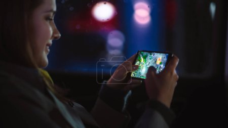 Glückliche Frau pendelt nachts auf dem Rücksitz eines Taxis nach Hause. Schöne Beifahrerin spielt Strategie-Videospiel auf Smartphone, während sie in einem Auto in der Stadtstraße mit arbeitenden Leuchtreklamen sitzt.