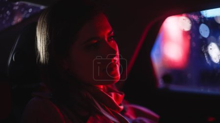 Foto de Primer plano Retrato de una mujer viajando a casa en un asiento trasero de un taxi por la noche. Hermosa pasajera mirando por la ventana mientras está en un coche en la calle de la ciudad con letreros de neón de trabajo. - Imagen libre de derechos