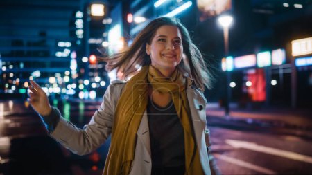 Téléchargez les photos : Portrait d'une belle femme heureuse en manteau de tranchée marchant dans une rue moderne avec des néons la nuit. Attrayant saut féminin autour du sourire, excité d'être dans un environnement cinématographique urbain. - en image libre de droit