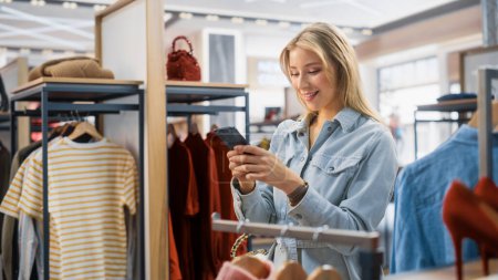 Photo for Beautiful Smiling Female Customer Shopping in Clothing Store, Using Smartphone, Browsing Online, Comparing on Internet, Choosing Stylish Clothes. Fashionable Shop, Colorful Brands, Sustainable Designs - Royalty Free Image