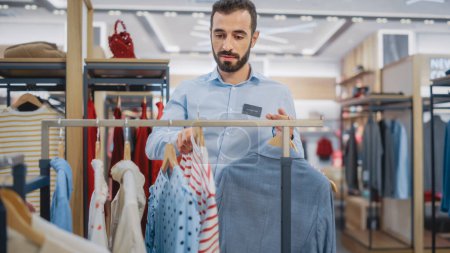Photo for Handsome Male Clothing Store Assistant Works in Fashionable Shopping Mall. Professional Shop Sales Retail Assistant Hangs New Colorful Collection with Sustainable Casual Design on Display Racks. - Royalty Free Image