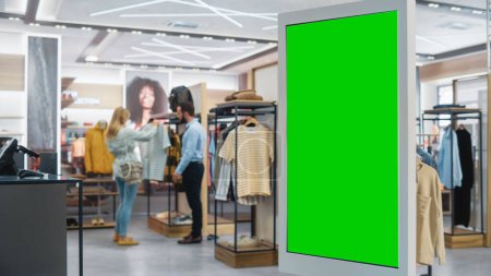 Téléchargez les photos : Prise de vue d'un écran tactile LCD au sol avec écran vert Chroma Key Mock Up debout dans le magasin de vêtements. Diverses personnes dans la boutique à la mode, Choisir et acheter des vêtements élégants. - en image libre de droit
