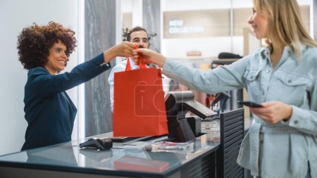 Photo for Clothing Store Checkout Cashier Counter: Woman and Male Retail Sales Managers Accept NFC Smartphone Payment from a Young Stylish Female Customer for Clothes and Pass a Recyclable Bag. - Royalty Free Image