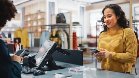 Photo for Clothing Store Checkout Cashier Counter: Woman Retail Sales Manager Accept NFC Smartphone and Credit Card Payments from a Young Female Customers for Clothes. - Royalty Free Image