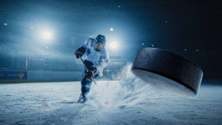Téléchargez les photos : Patinoire de hockey sur glace : Joueur professionnel tirant sur la rondelle avec bâton de hockey. Focus sur 3D Flying Puck avec effet Flou Motion. Prise de vue large dramatique, éclairage cinématographique. - en image libre de droit